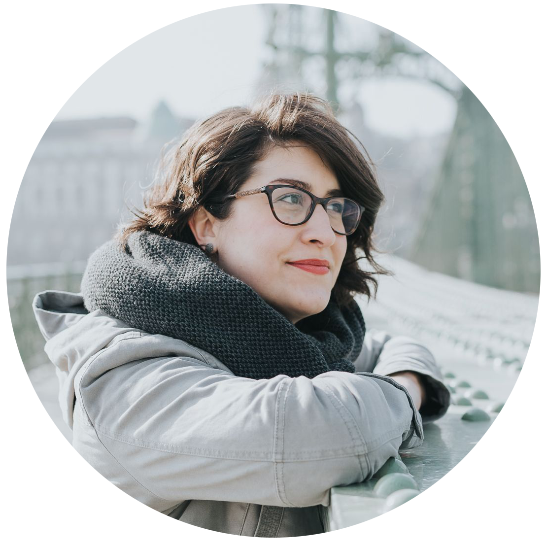 A woman with wavy brown hair and glasses wears a light grey coat and smiles while looking to the right